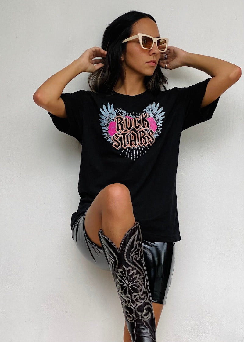 Camiseta negra de mujer con la frase "We just wanna be Rock Stars" en rosa, acompañada de alas celestes y rosas. Descubre más en Free Spirit.