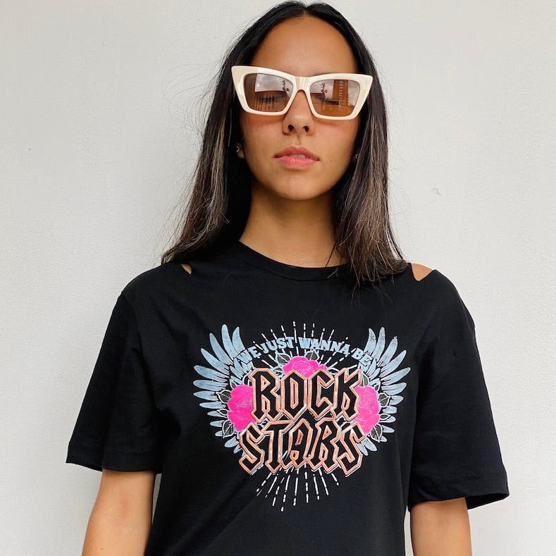 Camiseta negra de mujer con la frase "We just wanna be Rock Stars" en rosa, acompañada de alas celestes y rosas. Descubre más en Free Spirit.