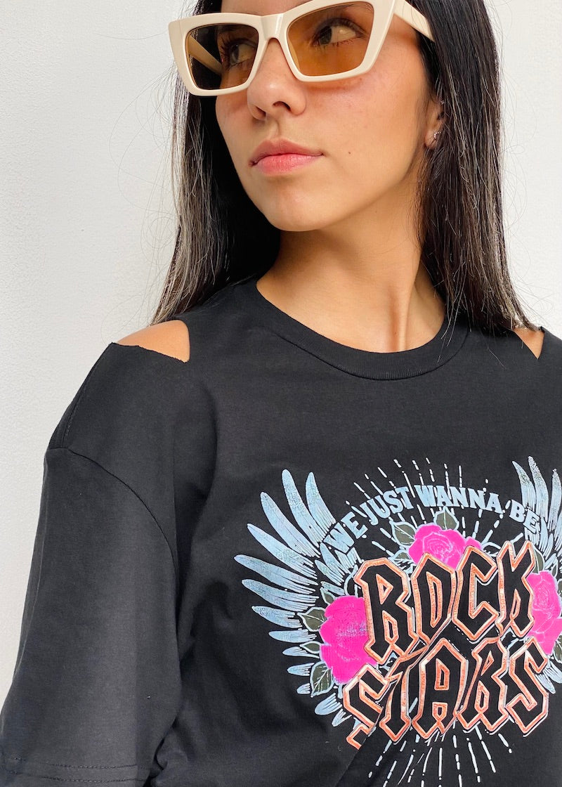 Camiseta negra de mujer con la frase "We just wanna be Rock Stars" en rosa, acompañada de alas celestes y rosas. Descubre más en Free Spirit.